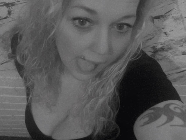 Close-up, grayscale photograph of JenniferCarmen with wet, wavy blonde hair, sticking out her tongue, and a nose piercing. She has a black top, showing cleavage, and a tattoo on her arm. JenniferCarmen's eyes are wide and she's looking directly at the camera, creating a sexy, playful vibe.