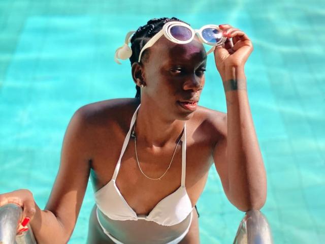 ElnaLethabo in een felblauw zwembad, met een witte bikini-top en een witte zonnebril op haar hoofd. Ze houdt een speelgoed voor het zwembad vast, haar natte huid glanst, en ze ziet er aantrekkelijk en geconcentreerd uit..