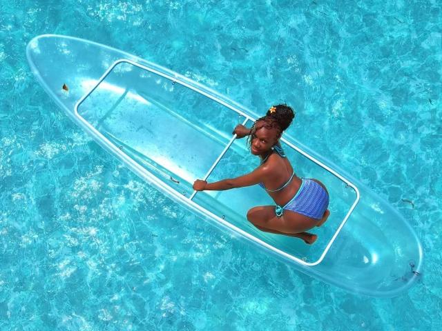 ElnaLethabo, uma mulher de pele escura com cabelos cacheados, ajoelha-se em um bote inflável azul brilhante em uma piscina azul clara. Ela veste um biquíni azul e branco xadrez. Sua expressão é divertida..