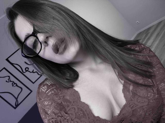 Photograph of BelleLovely, a white woman with long black hair, wearing black-framed glasses and a deep V-neck lace top that reveals ample cleavage. She looks down with a sultry expression.