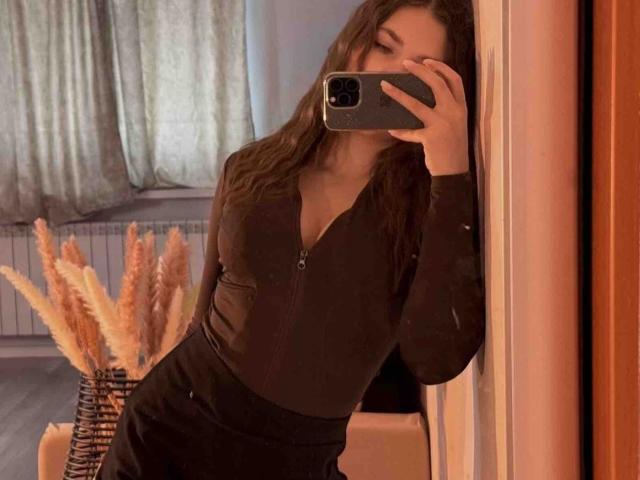 BelleLovely tirando uma selfie no espelho, vestindo uma blusa preta justa e calças pretas, segurando um celular com a mão esquerda, sua a mão direita apoiada em uma parede. Grama Pallas ao fundo.