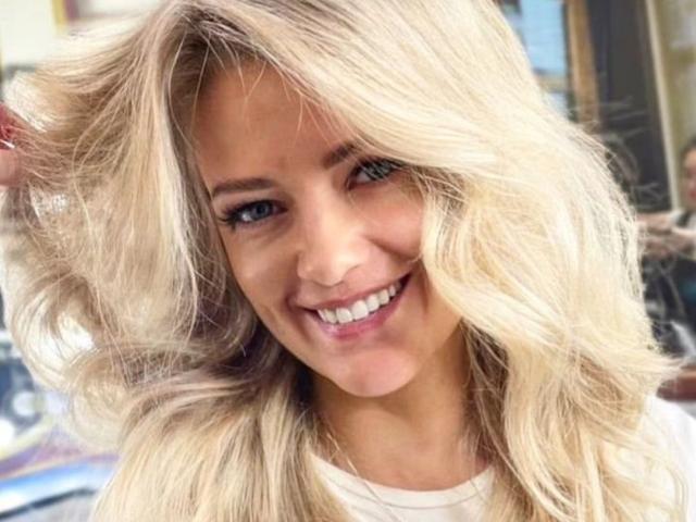 Close-up of GladisBelle, a blonde woman with wavy hair, smiling widely. She's touching her hair with her right hand, blue eyes, white shirt. Bright light highlights her features. Fucking sexy.