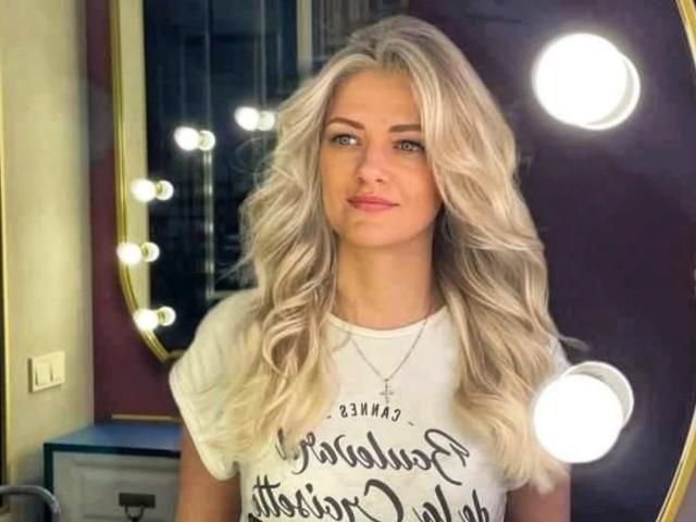 Photograph of GladisBelle with long, wavy blonde hair, wearing a white t-shirt with black text, standing in front of bright round lights. Sexy, confident pose, light makeup, blue eyes.