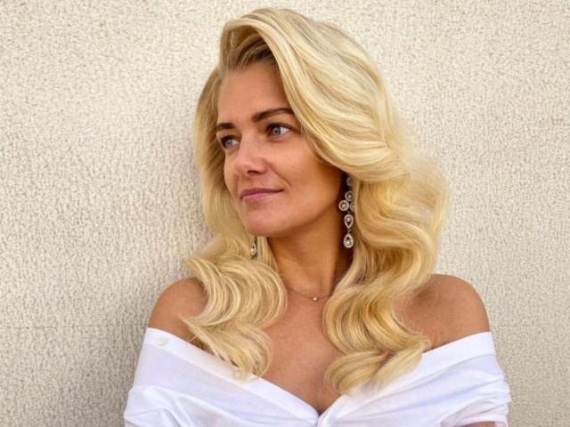 Photograph of GladisBelle, blonde woman with wavy hair, off-shoulder white top, diamond earrings, subtle smile, light skin, textured white wall background, subtle sexual tension, soft lighting, casual pose.