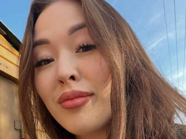 Close-up photo of MikeBooras with long brown hair, full lips with pink lipstick, and thick black eyelashes. Bright blue sky in background. Seductive expression, slight shadow on right cheek.