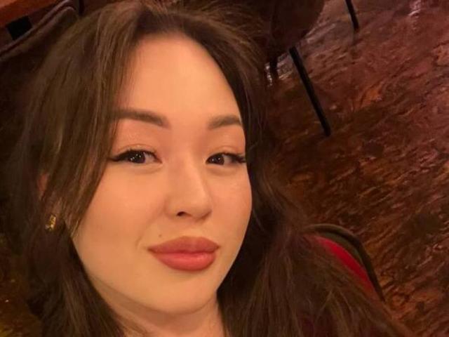 Close-up foto van MikeBooras met een lichte huid, bruin haar en rode lippenstift, met een lichte grijns. Zacht licht, vage bruine achtergrond. Lichte seksuele spanning, focus op het gezicht en de uitdrukking..