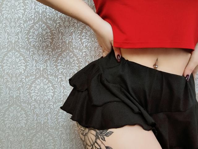 Close-up van het onderlichaam van Niferttiti, gekleed in een rode top en een zwarte rok, met een hand op de heup, zichtbare tatoeage op de dij en buik. Knop met gaatjes, donkere nagellak, gestructureerde witte achtergrond. Heel aantrekkelijk..