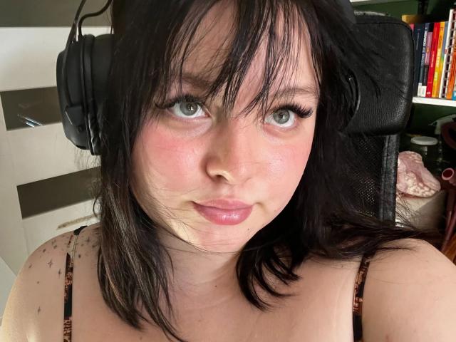 Close-up foto van Molly, een aantrekkelijke vrouw met zwart haar, blauwe ogen en roze wangen, die een koptelefoon draagt en een topje zonder bandjes aanheeft. Ze kijkt recht in de camera met een enigszins verleidelijke blik..