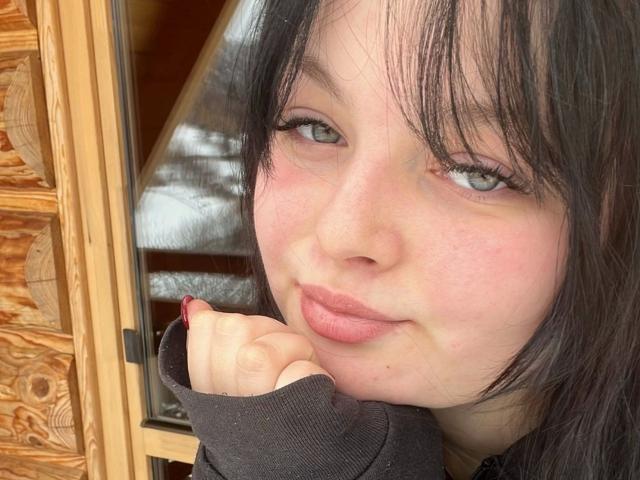Gros plan de MollyLolly-hot, une jeune femme à la peau pâle, aux cheveux noirs et aux yeux bleus, qui se repose le menton sur sa main. Portant un haut noir. Fond en panneaux de bois. Tellement mignon..