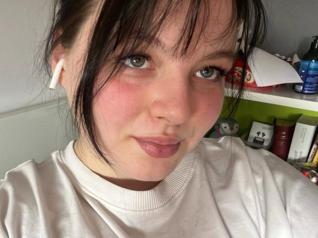 Primer plano de Molly. Una mujer atractiva, de piel clara, con cabello negro, flequillo y mejillas rosadas, que lleva una sudadera blanca. mirando directamente a la cámara con un ligero ceño fruncido, el fondo está lleno de artículos de tocador..