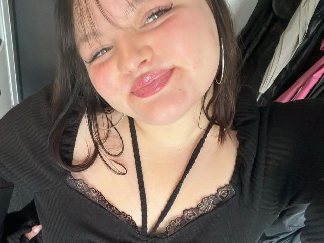 Close-up selfie of MollyLolly-hot with light skin, black hair, and red lipstick, wearing a black top with lace trim, showing cleavage, looking up with a slight smile, visible black strap.