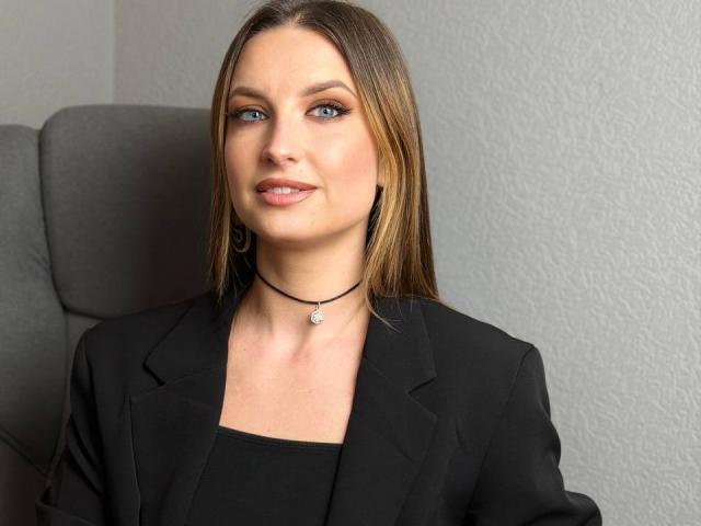Photograph of Milagres, a woman with straight brown hair, wearing a black blazer over a black top, and a black choker necklace, sitting in a grey office chair, looking slightly to the side with a subtle smile. Her blue eyes are slightly squinted. She has a slight, confident smirk.