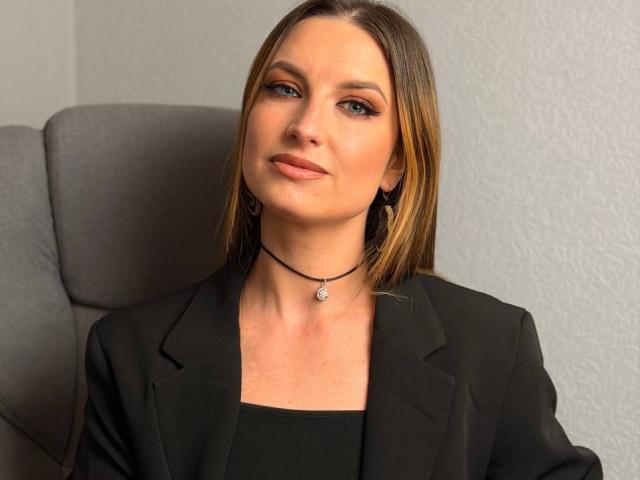 Fotografie von Milagres mit glattem, braunem Haar, die einen schwarzen Blazer und eine Choker-Kette trägt und in einem grauen Büro sitzt. Stuhl. Sie hat blaue Augen, ein leichtes Lächeln und einen selbstbewussten Blick..