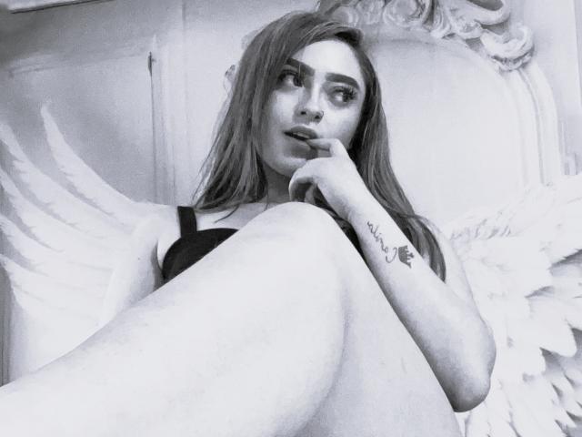 Photographie en noir et blanc de Samantha Rivera, avec de longs cheveux, assise les jambes relevées, la main près de la bouche, portant un haut noir. tatouage au poignet, pose suggestive, textures évoquant des ailes en arrière-plan.