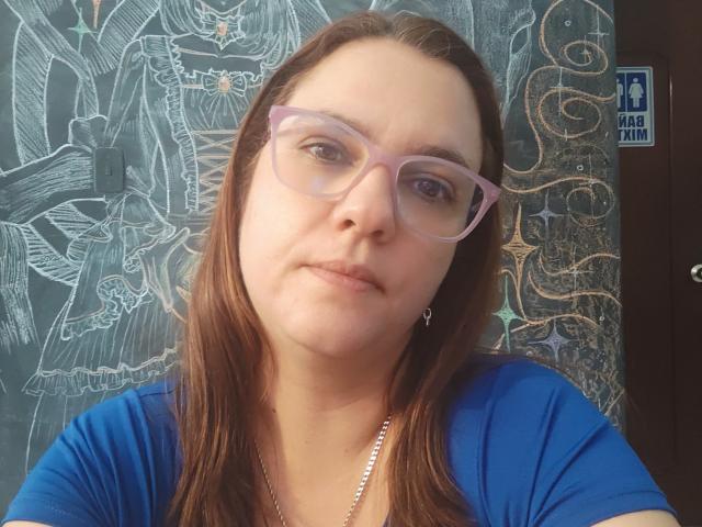 Amelia Wilson, de piel clara y cabello castaño, que lleva gafas rosas y una camisa azul, está de pie frente a un objeto detallado, pared con diseño. Ella está mirando directamente a la cámara, con una expresión sutil y segura..