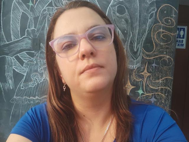 Amelia Wilson, con capelli marrone medio, indossa occhiali viola e una camicia blu, è in piedi di fronte a un muro nero con delle parti bianche. Disegni astratti.
ha lo sguardo leggermente rivolto verso l'alto, con un'espressione neutra..
