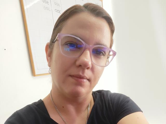 Photograph of AmeliaWilson with brown hair in a ponytail, wearing purple glasses and a black t-shirt, looking directly at the camera with a slight smirk. She has a silver necklace.