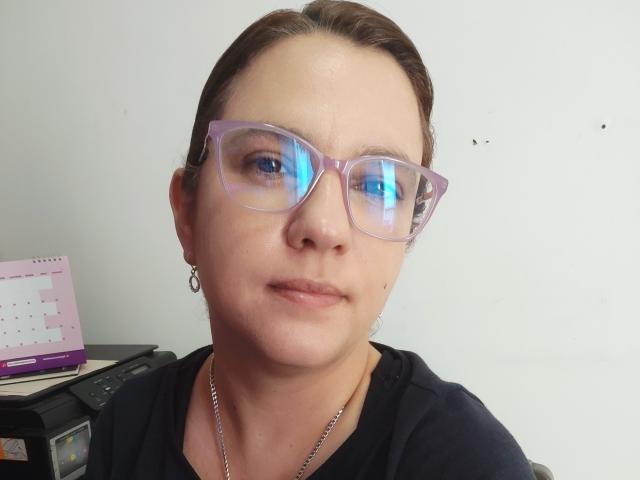 AmeliaWilson has fair skin and brown hair pulled back, wearing clear, reflective glasses and a black top. She's looking directly at the camera, with a laptop in the background. Fucking serious look.