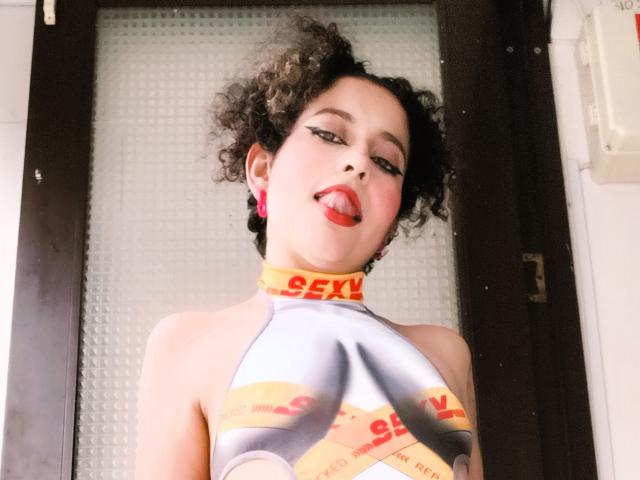 Close-up foto van IrinaSweett, met krullend zwart haar, die haar tong uitsteekt op een uitdagende manier. Het draagt een glanzend zilveren topje met een rode "SEX"-halsband, waardoor de kleine borsten worden benadrukt. Hel licht, achtergrond met een getextureerde witte kleur..