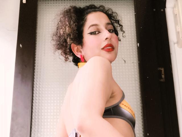 IrinaSweett, une femme aux formes généreuses, avec des cheveux noirs bouclés, du rouge à lèvres et un soutien-gorge de sport noir, jette un coup d'œil par-dessus son épaule. Son regard était séduisant, son bras levé, dévoilant son côté. Fond clair.