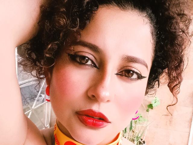 Fotografia ravvicinata di IrinaSweett, con capelli scuri e ricci, rossetto rosso e un audace eyeliner nero. Lei guarda direttamente la fotocamera con un'espressione leggermente seducente. Una luce intensa illumina il suo viso.