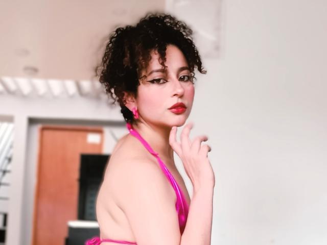 Digitale foto van IrinaSweett, een mollige, lichtgetinte vrouw met kort, krullend zwart haar, die een felroze top met halterneck en een rode lipstift, hand op kin, verleidelijk over de schouder kijkend, helder witte achtergrond.