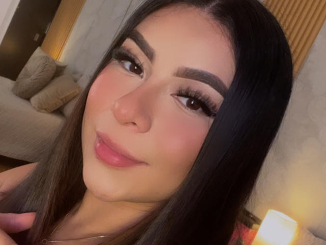 Close-up foto van GabrielaFerrer, een jonge vrouw met lang zwart haar, dikke wenkbrauwen en volle, roze lippen. Ze heeft een subtiele glimlach en kijkt recht in de camera. Warme verlichting.