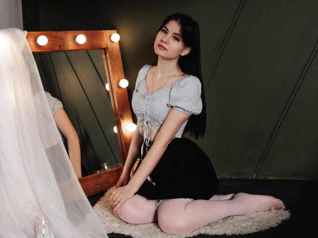 ChrissieCandy kneels on a white shag rug, wearing a light blue, puffed-sleeve top and black skirt, with white stockings, in front of a mirror with lit bulbs. Fucking sexy.