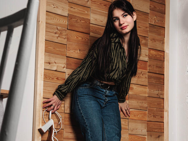 ChrissieCandy, une femme séduisante avec de longs cheveux noirs, est appuyée contre un mur en bois, vêtue d'une chemise rayée verte et moulante. jeans bleus.
elle tient des câbles blancs dans sa main droite.