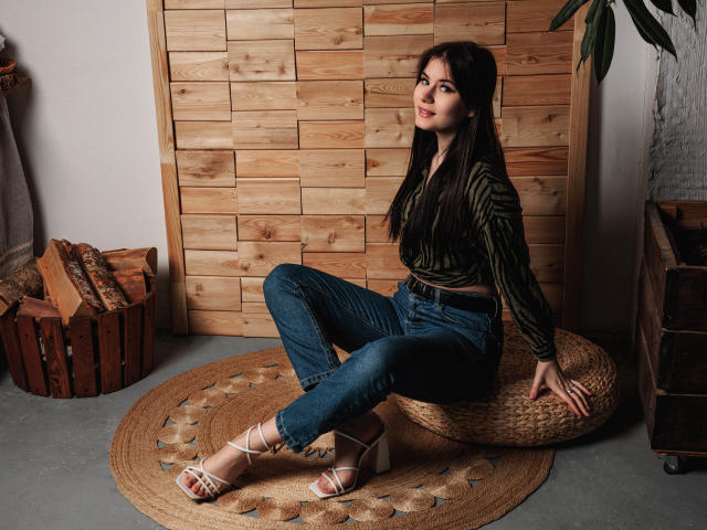 ChrissieCandy, uma morena atraente com cabelos longos, está sentada em um tapete trançado, com as pernas cruzadas, vestindo jeans justos e sandálias finas. sandálias, olhando para trás de forma sedutora. Fundo de painel de madeira, banquinho de madeira. Incrivelmente atraente..