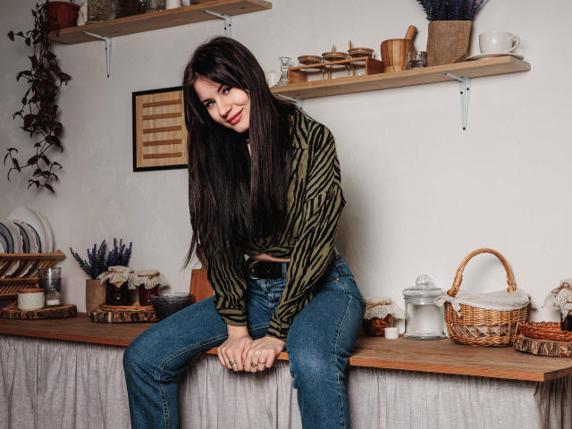 ChrissieCandy, con i suoi lunghi capelli neri, è piegata in avanti su un bancone di legno, indossa una camicia a righe verdi e jeans blu. mani sulle ginocchia, cesti e ceramiche sugli scaffali, sfondo di parete bianca.