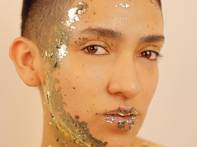 Close-up of MichaelaMistress with shaved head, golden glitter covering one side of her face and neck, brown eyes, neutral expression, soft skin, brown lips, bright light, stark contrast.