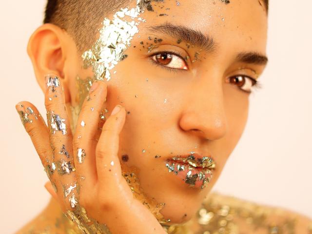 Close-up van MichaelaMistress, een persoon met een lichte huidskleur en kort haar, bedekt met glitters. MichaelaMistress raakt hun wang aan met een hand en kijkt recht in de camera met bruine ogen. Glitter op gezicht en hand
.