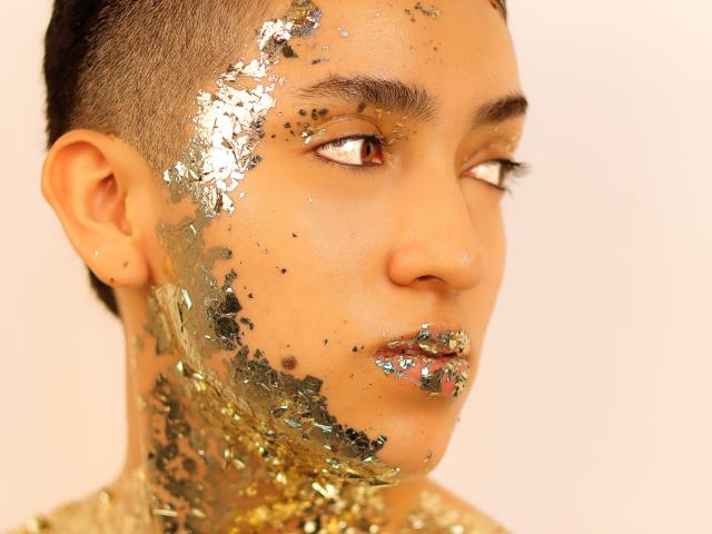 Close-up van MichaelaMistress, met metallic goud- en zilverglitters die de linkerhelft van haar gezicht en nek bedekken. Ze kijkt naar rechts met halfgesloten ogen, met kort zwart haar. Heel erg aantrekkelijk..