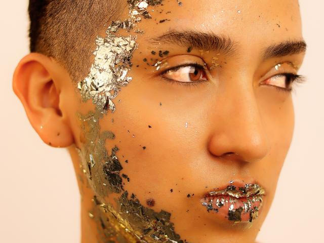 Gros plan du visage de MichaelaMistress, avec un maquillage très pailleté du côté gauche. Ses yeux regardent de côté, ses lèvres sont légèrement entrouvertes et ornées de paillettes. De fines paillettes dorées et noires recouvrent sa joue. Un éclairage vif.
.