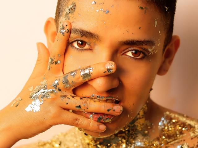 Close-up photo of MichaelaMistress with dark skin, brown eyes, and short hair, covering her mouth with a hand covered in glitter. Shiny glitter on her face and shoulders, intense gaze.