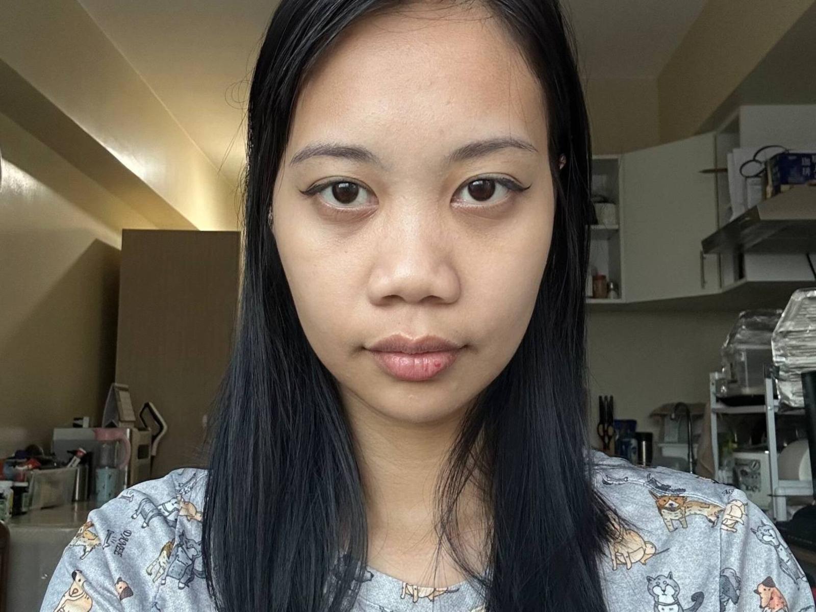 GloryGlory tem cabelo preto liso, pele marrom clara e uma expressão neutra. Vestindo uma camisa floral azul clara. O fundo mostra itens de cozinha, incluindo prateleiras e recipientes. Close-up com foco, tensão sexual sutil..