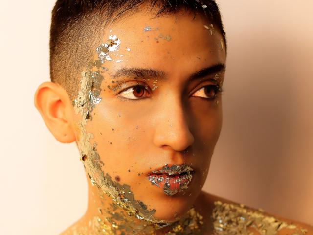 Close-up de MichaelaMistress, um jovem com cabelo preto curto, coberto de glitter no rosto e no pescoço. Ele olha fixamente para o lado, com seus intensos olhos castanhos. A iluminação forte realça o brilho..