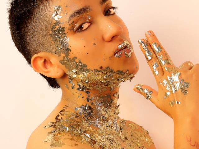 Close-up of MichaelaMistress, an attractive man with short black hair, orange skin, and brown eyes. His face and hand covered in glittery gold flakes, finger up in a seductive gesture.