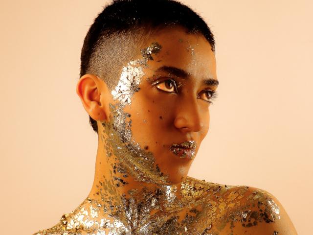 Close-up of MichaelaMistress, a young man with short black hair, glistening wet skin, intense brown eyes, and a sultry gaze, partially covered in glittery, reflective particles. Golden light highlights his muscular shoulders.