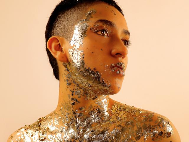 Photographie d'un gros plan, en profil, de MichaelaMistress, avec des cheveux courts et foncés, et une texture de peau brillante et scintillante. visage et épaules, éclairage jaune doré, regard intense, expression neutre, aucun vêtement visible..