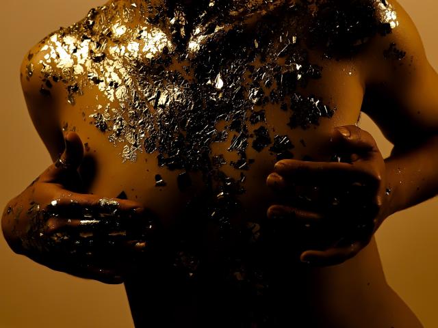 Close-up of a nude MichaelaMistress with a shiny, glittery body covering her chest. She's pressing her large, round breasts with her hands, creating a provocative, sexual tension. Golden light highlights her curves.