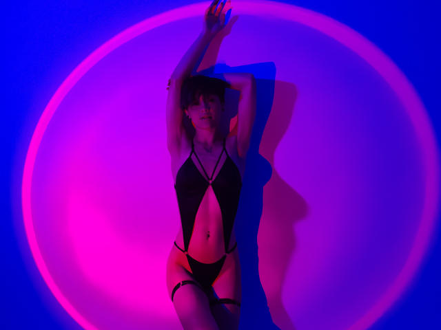 LissyLuxe in zwarte, elegante lingerie, met armen omhoog, verlicht door een roze en blauwe neoncirkel. Ze is aantrekkelijk, met een subtiele grijns, en straalt uit tegen de levendige lichten. Duidelijke, erotische sfeer.