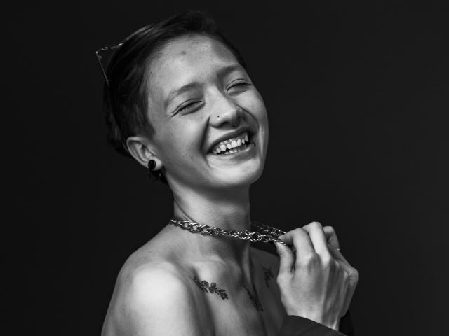 Black and white photo of LissyLuxe with short hair, smiling widely, holding a chain necklace with right hand, tattoos on chest, dark background. High contrast, sexy, confident vibe.