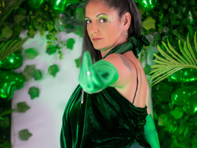 Madame Scarlett, con cabello negro, maquillaje de ojos verde y un vestido de terciopelo negro, extiende su mano verde brillante hacia adelante. Fondo de color verde con hojas y hexágonos. Mirada sensual e intensa, aura brillante..