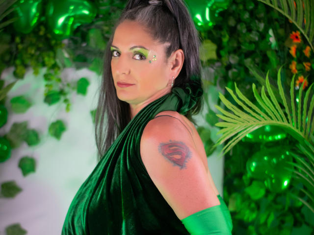 Madame Scarlett, donna dai capelli scuri con trucco verde per gli occhi, sciarpa verde, tatuaggio sulla spalla destra, sfondo illuminato di verde, piante verdi e rigogliose, che guarda indietro per spalla, sguardo intenso, colori vivaci, atmosfera sensuale..