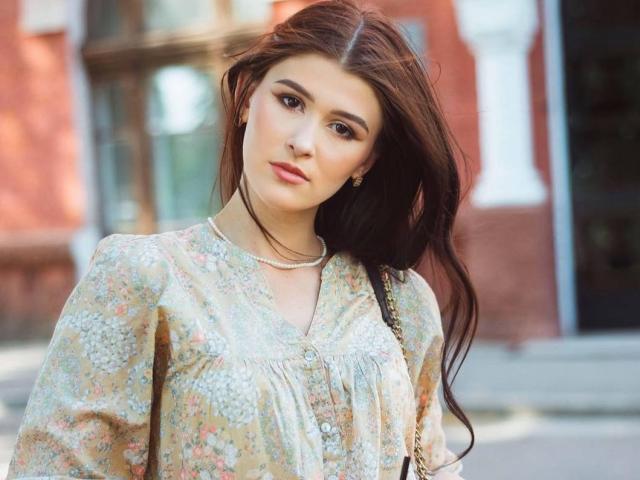 Fotografie von AngelSmooth, einer jungen Frau mit langen, dunkelbraunen Haaren, die eine Bluse mit Blumenmuster und weißer Spitze trägt. schneiden. Sie steht in einem von der Sonne beschienenen Stadtgebiet und blickt direkt in die Kamera, mit einem leichten Lächeln. Ihr Gesichtsausdruck ist entspannt, mit einem Hauch von sexueller Spannung. Im Hintergrund sind unscharfe, rote Backsteingebäude zu sehen..