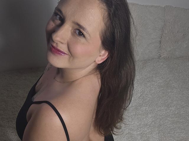 Photograph of JoyDiamond, a smiling woman with fair skin and dark brown hair, wearing a black strapless top, against a textured gray wall. Her red lips and slight blush add allure.