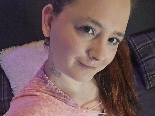 Photo en gros plan de JoyDiamond, une femme à la peau claire avec des cheveux bruns, portant un sweat à capuche rose avec un col en fourrure blanche. léger sourire, petit tatouage sur le cou, fond sombre, boucle d'oreille visible..