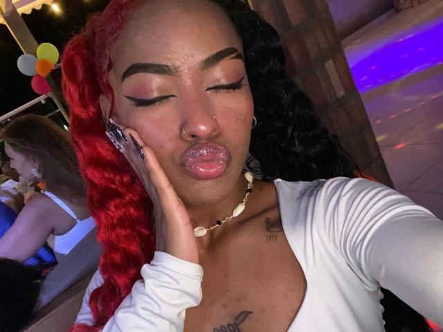 Nahaufnahme von SummerWilliams mit halblanger, rot-schwarzer Haare, geschlossenen Augen, leicht geschürzten Lippen, goldenem Nasenring, weißem Oberteil. Hand vor dem Gesicht, Neonlichter, Tattoos auf der Brust, sexy, lebendige Farben..